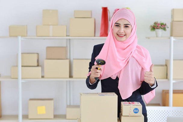 Active asian woman wearing muslim suit standing and using scanner and showing thumb up with package box delivery. Startup small business SME freelance girl work at home concept - Photo, Image