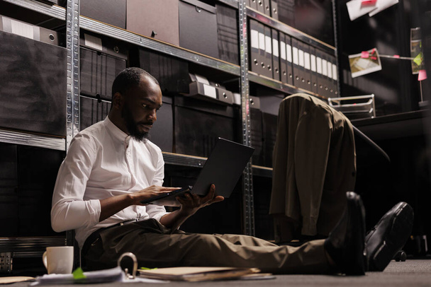 Afrykański policjant siedzący na podłodze i korzystający z laptopa. Kryminolog badający akta sprawy kryminalnej, sprawdzający informacje na komputerze przenośnym w ciemnym biurze w nocy - Zdjęcie, obraz