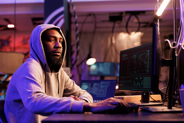 Hacker v kapuci hackuje počítačový systém a dívá se do kamery. Mladý africký Američan porušuje zákon a porušuje data, zatímco pracuje pozdě v opuštěném skladu portrét - Fotografie, Obrázek