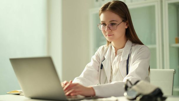 Skupiony lekarz siedzi przy stole w szpitalu i pracuje na laptopie online. Skoncentrowany pielęgniarka lub terapeuta patrząc na ekranie komputera i konsultacji pacjenta na gadżet - Zdjęcie, obraz