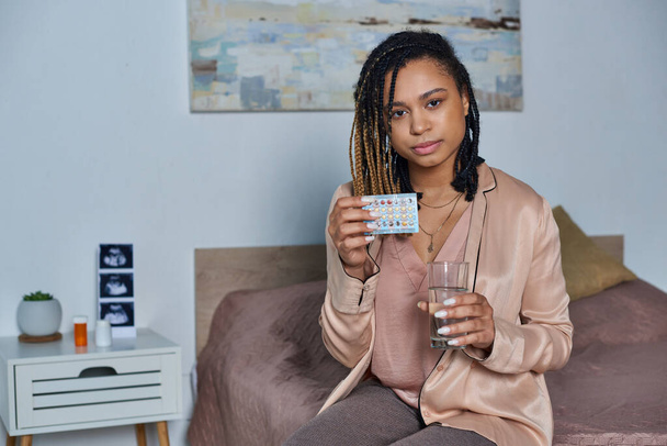 Afrikai amerikai nő kezében pohár víz és fogamzásgátló tabletták, terhesség, modern hálószoba - Fotó, kép