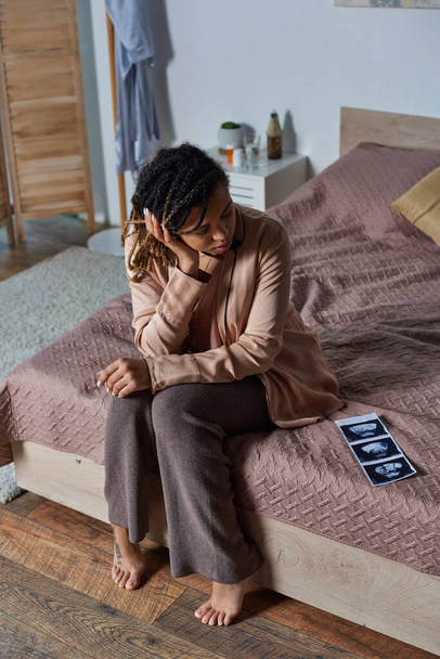 Αφροαμερικανή που κάθεται στο κρεβάτι και κοιτάει φωτογραφίες υπερήχων, ένα ποτήρι νερό, αντισύλληψη. - Φωτογραφία, εικόνα