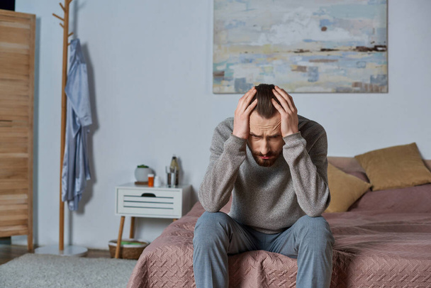 gestresste man wegkijkend en zittend op bed in moderne slaapkamer, schilderen op muur, eenzaamheid, bezorgdheid - Foto, afbeelding