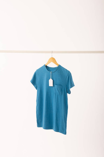 Blue t shirt with tag on hanger hanging from clothes rail with copy space on white background. Fashion, clothes, colour and fabric concept. - Photo, image
