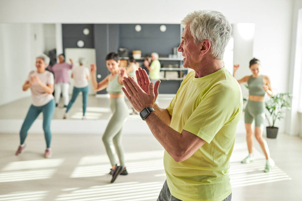 Hombre mayor bailando durante el entrenamiento deportivo con otras personas de fondo - Foto, imagen