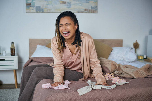 wanhoop, Afro-Amerikaanse vrouw huilt in de buurt van baby kleding op bed, miskraam concept, depressie - Foto, afbeelding
