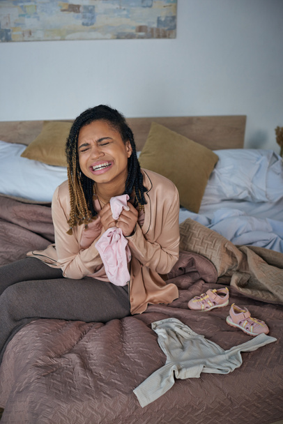 Trauer, afrikanisch-amerikanische Frau weint in der Nähe von Babykleidung auf dem Bett, Fehlgeburtenkonzept, Depression - Foto, Bild