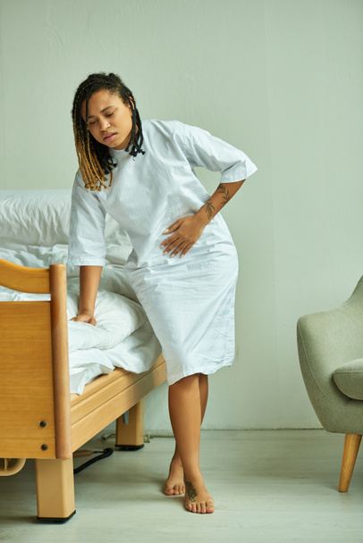 femme afro-américaine tatouée en robe d'hôpital touchant le ventre, salle privée, patient à la clinique - Photo, image