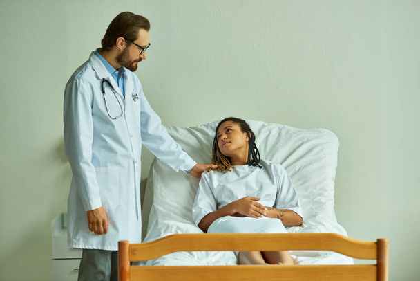 dokter in bril staan in de buurt van Afrikaans-Amerikaanse vrouw in ziekenhuis jurk, prive-afdeling, patiënt - Foto, afbeelding
