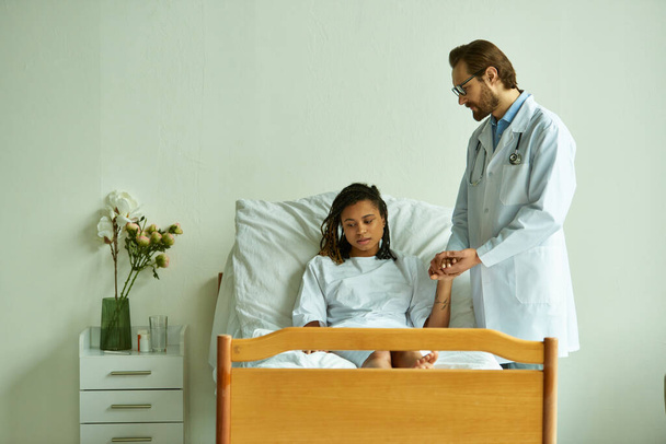 vousatý lékař v brýlích drží ruku africké Američanky v nemocničním rouchu, soukromé oddělení - Fotografie, Obrázek