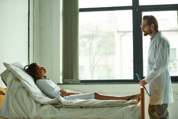 doctor de pie con portapapeles cerca de la mujer afroamericana, sala privada, hospital, consulta - Foto, imagen