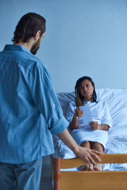 marido de pie cerca de sala privada de triste esposa afroamericana, hospital, concepto de aborto involuntario - Foto, imagen
