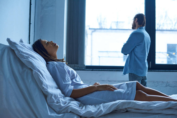 Концепция выкидыша, депрессивная африканская женщина скорбит, лежит в больничной койке рядом с мужем - Фото, изображение