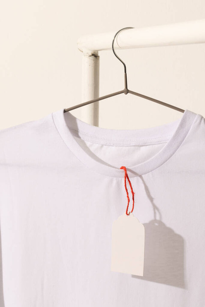 White t shirt with tag hanging from clothes rack with copy space on white background. Fashion, clothes, colour and fabric concept. - Фото, изображение