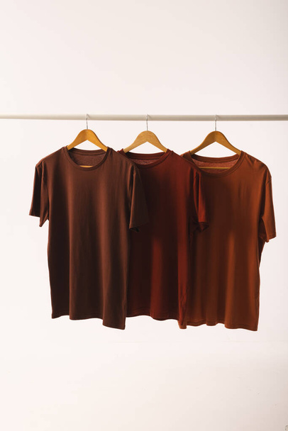 Drei braune T-Shirts auf Kleiderbügeln hängen an der Kleiderstange und kopieren Platz auf weißem Hintergrund. Mode, Kleidung, Farb- und Stoffkonzept. - Foto, Bild