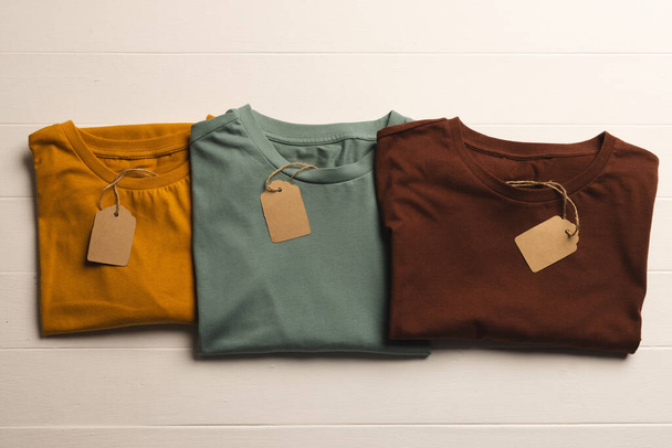 Close up of flat lay of three t shirts with tags and copy space on white boards background. Fashion, clothes, colour and fabric concept. - Foto, Bild