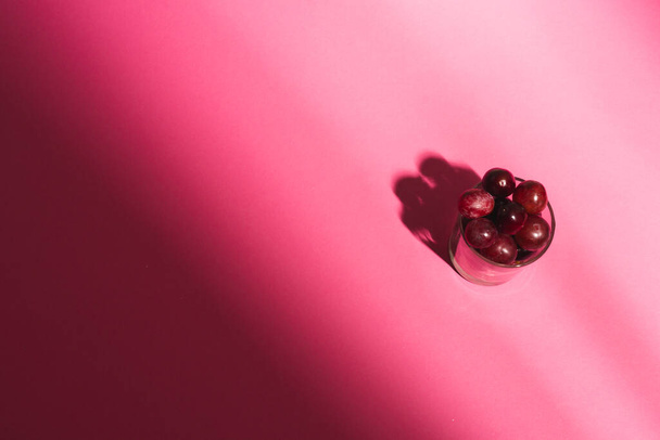 Κοντινό πλάνο του γυαλιού των κόκκινων σταφυλιών και αντίγραφο χώρο σε ροζ φόντο. Φρούτα, τρόφιμα, φρεσκάδα και χρωματική έννοια. - Φωτογραφία, εικόνα