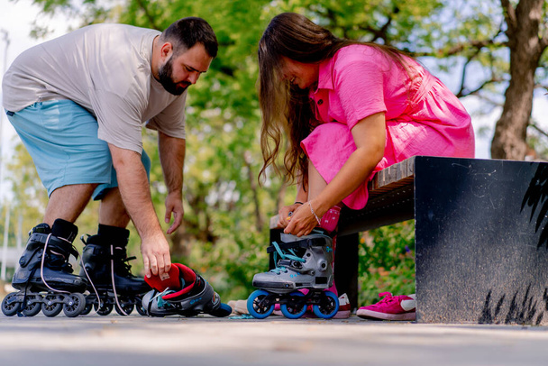 Young couple guy helps girl put on rollerblades who is sitting on a bench in the park while dating plus size models - Photo, Image