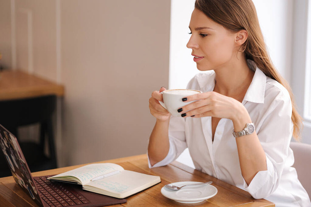 Młoda kobieta pije kawę w kawiarni. Kobieta w białej koszuli trzymać filiżankę cappuccino i uśmiech. Formalny portret. Wysokiej jakości zdjęcie - Zdjęcie, obraz