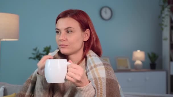 retrato de mujer joven envuelta en una manta caliente toma un sorbo de té caliente sosteniendo la taza en las manos y tratando de mantener el calor, mientras está sentado en el sofá en la habitación fría - Imágenes, Vídeo