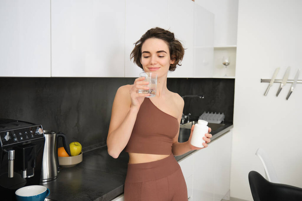 Portrét fitness dívky, zdravé ženy stojící v kuchyni s prášky, bere vitamíny se sklenicí vody pro zdravou pohodu. Sportovkyně drží doplňky stravy. - Fotografie, Obrázek