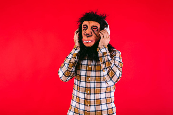 チンパンジーの猿のマスクとチェッカーのシャツを持つ男,赤い背景でホワイトヘッドホンで音楽を聞く. - 写真・画像
