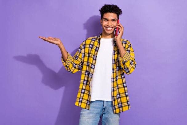 Portrait of toothy beaming man wear plaid shirt talking on smartphone palm demonstrate product isolated on violet color background. - Фото, зображення