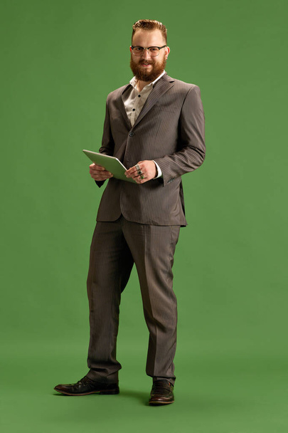 Full-length portrait of bearded stylish businessman in suit and glasses posing with tablet, looking at camera against green background. Concept of business, emotions, lifestyle, office, fashion, ad - Foto, imagen
