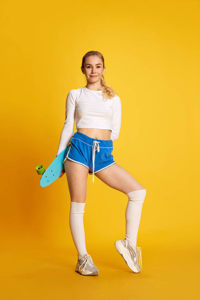 Full lenght shoot of smiling young girl wearing sportswear with ponyboard over yellow background. Exercises. Concept of sport, hobby, fitness, fashion, human emotions, beauty, healthy lifestyle, ad - Photo, Image
