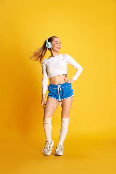 Vertical full lenght photo of young smiling sporty woman with headphones looking away and over yellow background. Concept of sport, hobby, fitness, healthy lifestyle, ad - Photo, Image