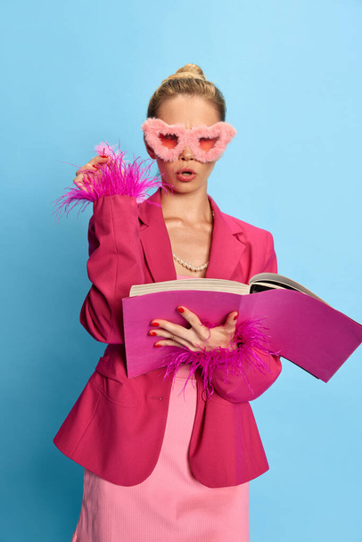Ritratto di ragazza cahrming, donna che indossa glamour in occhiali insoliti, tenendo il libro di testo guardando la fotocamera su sfondo azzurro studio. Concetto di moda, bellezza, emozioni positive, business.Ad - Foto, immagini