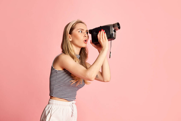 Woman dressed stylishly looking at handheld vintage camera against light pink background. Concept of job, filming, lifestyle and ad - Photo, image