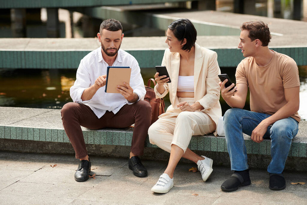 Συνάδελφοι ξεκουράζονται έξω κατά τη διάρκεια του διαλείμματος, μιλώντας και ελέγχοντας τα μέσα κοινωνικής δικτύωσης σε smartphones και tablets - Φωτογραφία, εικόνα