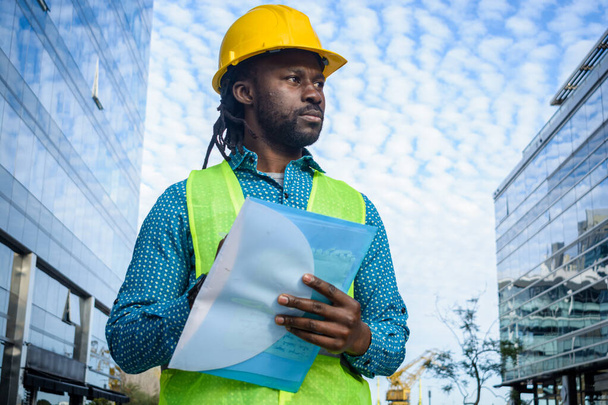 portrait of black man with beard and dreadlocks, architect in yellow safety helmet and vest, standing outdoors thinking holding a folder, copy space. - Photo, Image