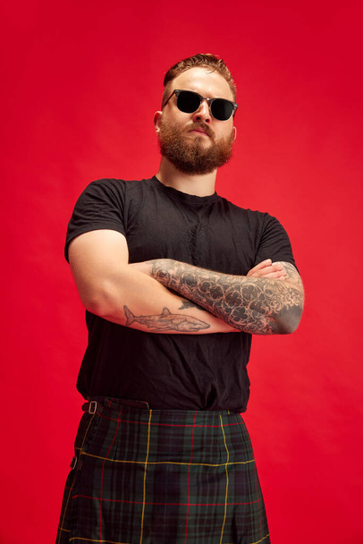 Portrait of bearded serious man in sunglasses posing in scottish skirt - kilt against red studio background. Extraordinary look. Concept of lifestyle, scottish style, fashion, music, fun and joy, ad - Photo, image