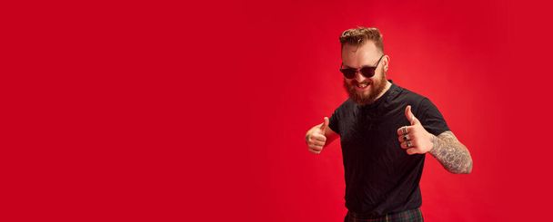Portrait of bearded stylish man in sunglasses cheerfully posing against red studio background. Concept of lifestyle, scottish style, fashion, music, fun and joy. Banner. Copy space for ad - Zdjęcie, obraz