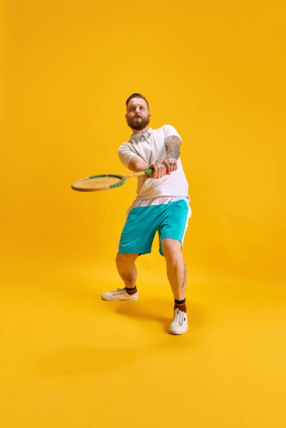 Full-length image of bearded man in sportswear posing with tennis racket against yellow studio background. Concentration, game. Concept of sport, strength, fashion, emotions, lifestyle, ad - Photo, Image