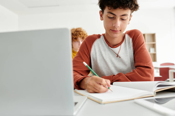 adolescent écolier écriture sur notebook près ordinateur portable et flou camarade de classe dans salle de classe à l'école - Photo, image