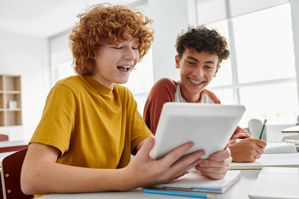 Pozitivní dospívající školák pomocí digitálního tabletu s přítelem během výuky ve třídě na pozadí - Fotografie, Obrázek