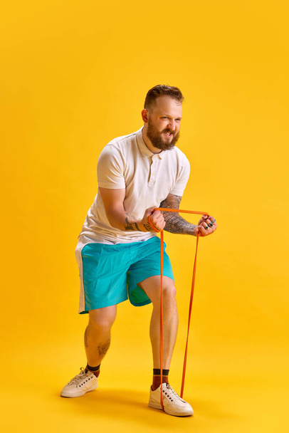 Full-length image of bearded man in sportswear training in sportswear against yellow studio background. Workout. Concept of sport, strength, fashion, emotions, lifestyle, ad - Foto, imagen