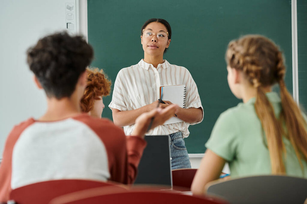Pensive african american teacher holding notebook near pupils and laptop in classroom - Foto, imagen