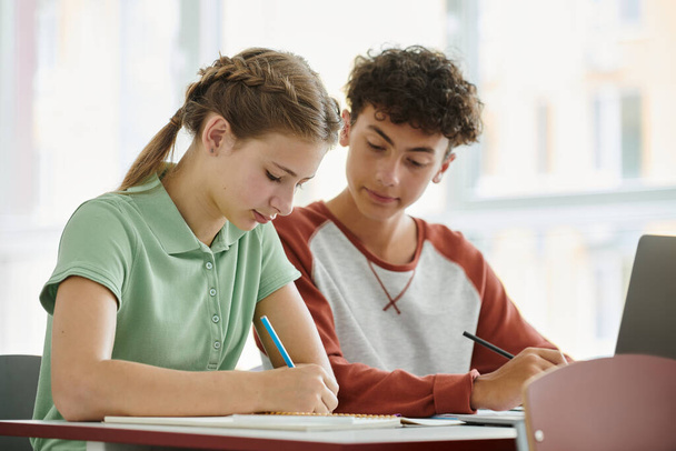 Adolescente escolar escribiendo y mirando cuaderno cerca de compañero de clase durante la lección en el aula en la escuela - Foto, imagen