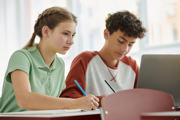 Підліток школярка пише на блокноті біля розмитого однокласника і ноутбука під час уроку в класі - Фото, зображення