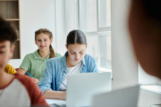 Teenager Schulmädchen schaut in die Kamera und lächelt in der Nähe verschwommener Klassenkameraden während des Unterrichts im Klassenzimmer - Foto, Bild
