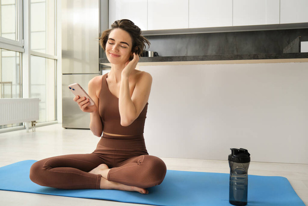 Bild einer sportlichen Frau in Aktivkleidung, sitzt zu Hause auf einer Yogamatte, hält das Smartphone in der Hand, nutzt Workout-App, sucht online nach Übungen. Kopierraum - Foto, Bild