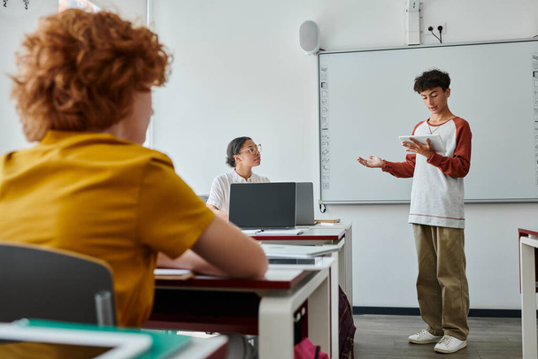 Teenage schoolboy talking while holding digital tablet near african american teacher in classroom - Foto, Bild