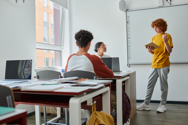 Vörös hajú tini iskolás gazdaság könyv és beszél közelében afro-amerikai tanár tanítás során az osztályban - Fotó, kép