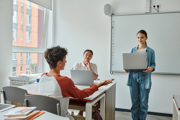 Adolescente colegial hablando con compañero de clase con portátil cerca de profesor afroamericano en el aula - Foto, Imagen