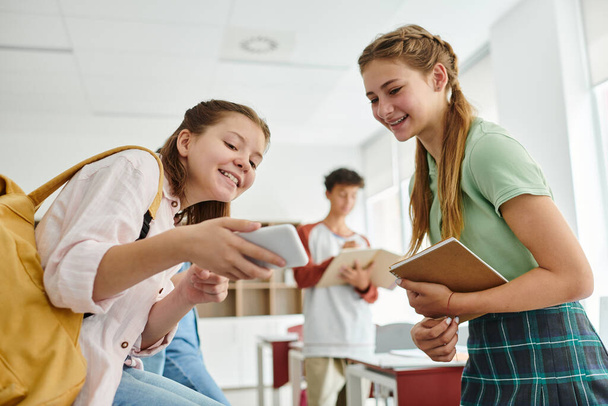 Lächelnde Teenie-Schulmädchen mit Rucksack und Notizbuch mit Smartphone im Klassenzimmer - Foto, Bild