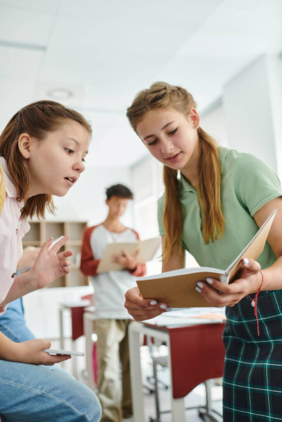 Colegiala adolescente mostrando cuaderno a compañero de clase con teléfono inteligente en el aula borrosa en la escuela - Foto, Imagen